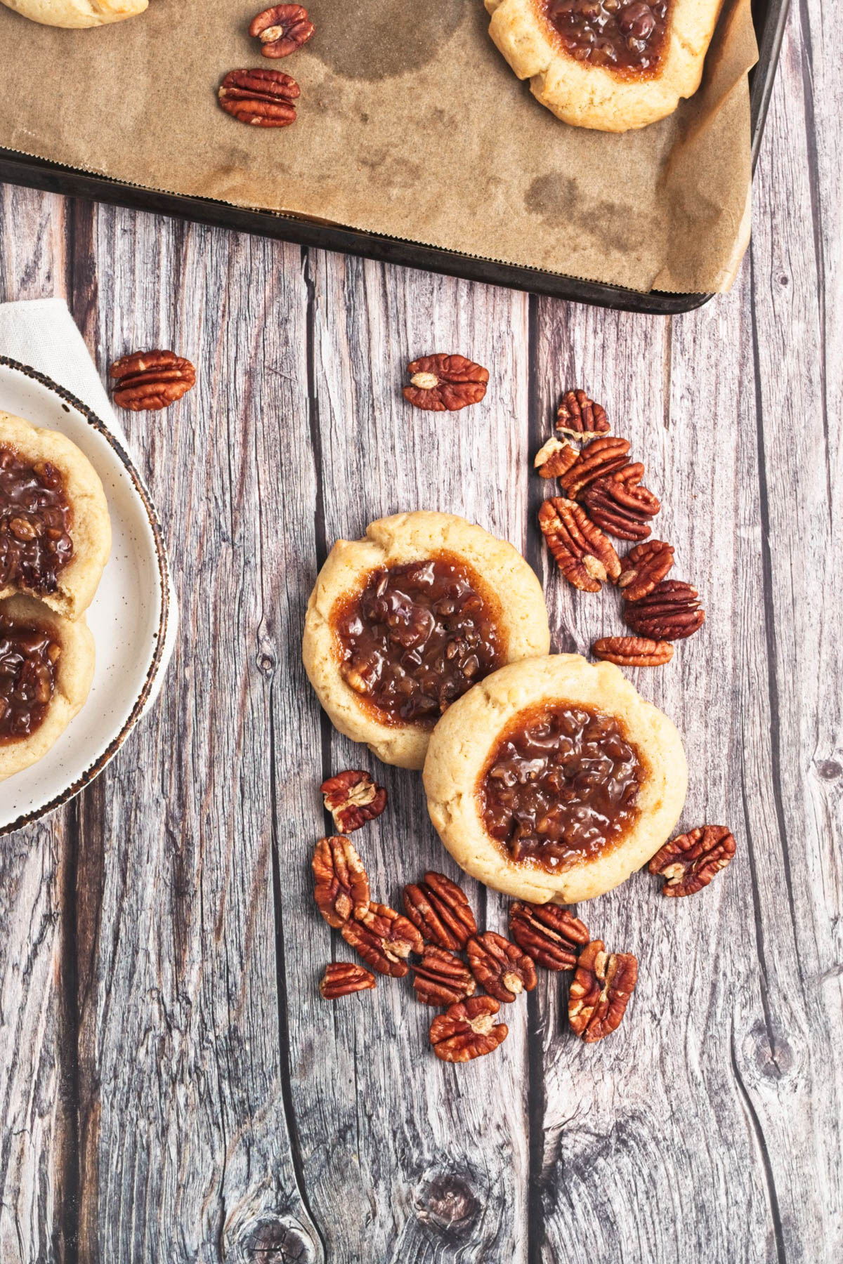 Two pecan pie cookies on a wood board with pecans scattered and a baking sheet of cookies in background.