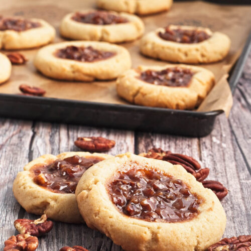 Two pecan pie cookies on a wood board with pecans scattered and a baking sheet of cookies in background.