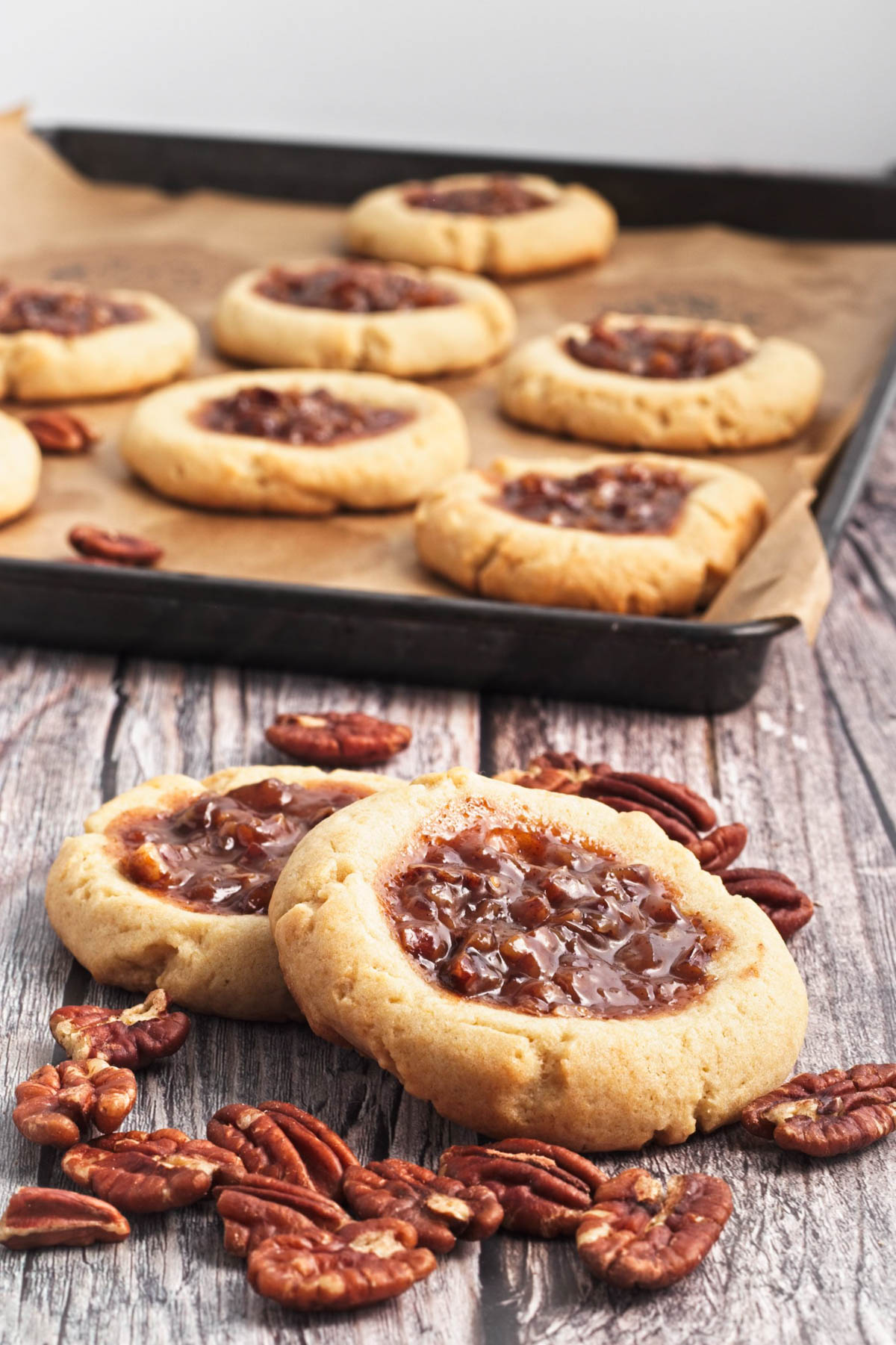 Two pecan pie cookies on a wood board with pecans scattered and a baking sheet of cookies in background.