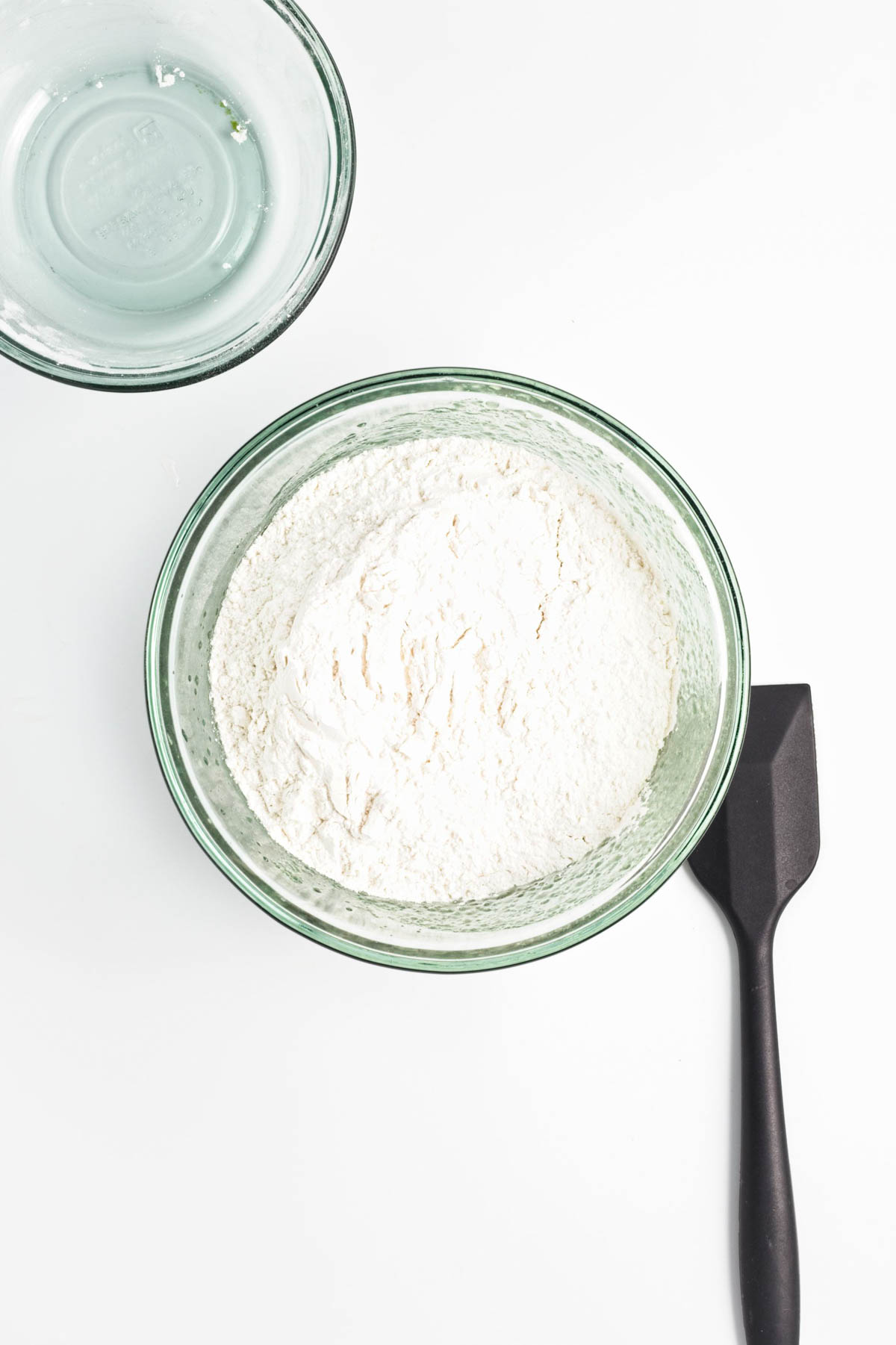 Glass bowl of flour with black spatula and empty glass bowl above.