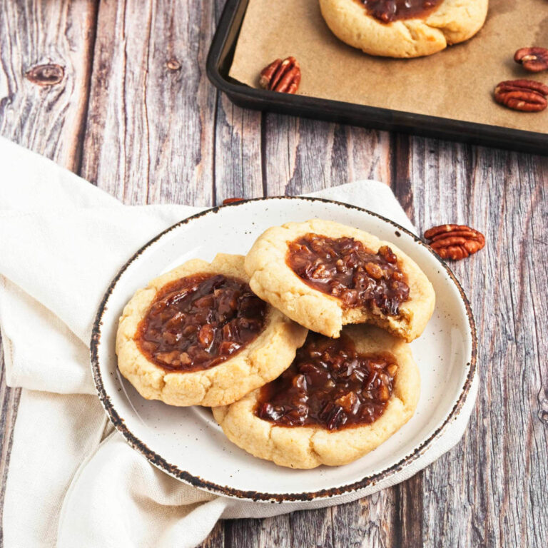 Pecan pie cookies on plate with more in the back on cookie sheet on wooden table.