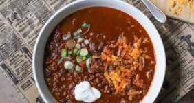 Two white bowls filled with chili and topped with spring onions, sour creme, and cheese on a newspaper with small bowls of green onions, sour cream, and a block of cheese with shredded cheese.