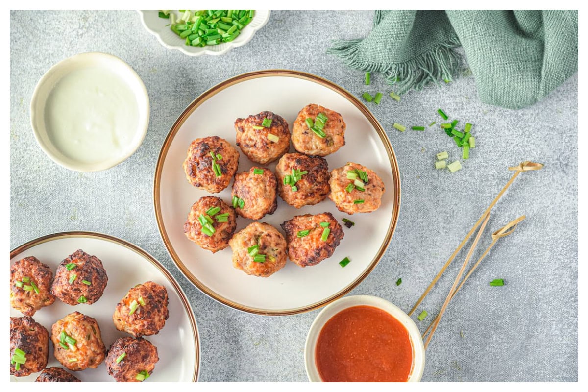 Meatballs on a white plate with a bowl of hot sauce and green onions on the side. 