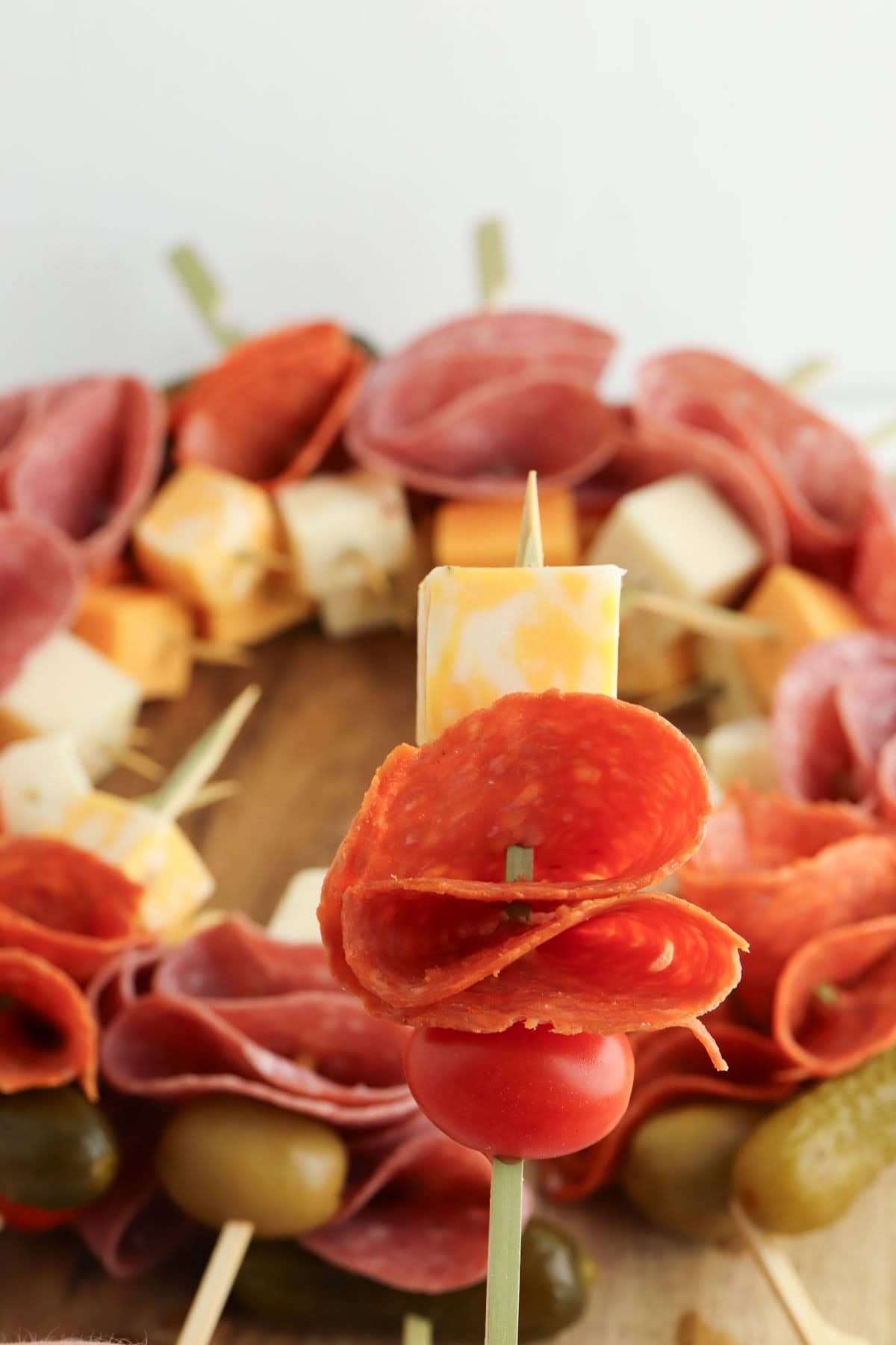 Tomato, pepperoni, and a cube of cheese on a skewer with charcuterie wreath in the background.