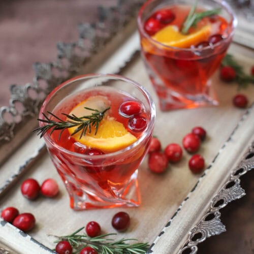 Red cocktail in a lowball glass with rosemary, cranberries, and an orange slice on a tray.