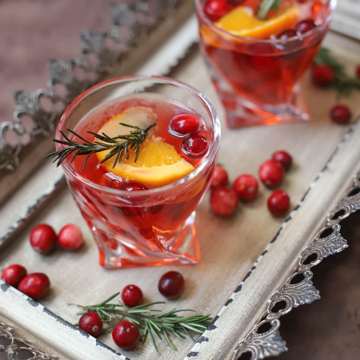 Red cocktail in a lowball glass with rosemary, cranberries, and an orange slice on a tray.