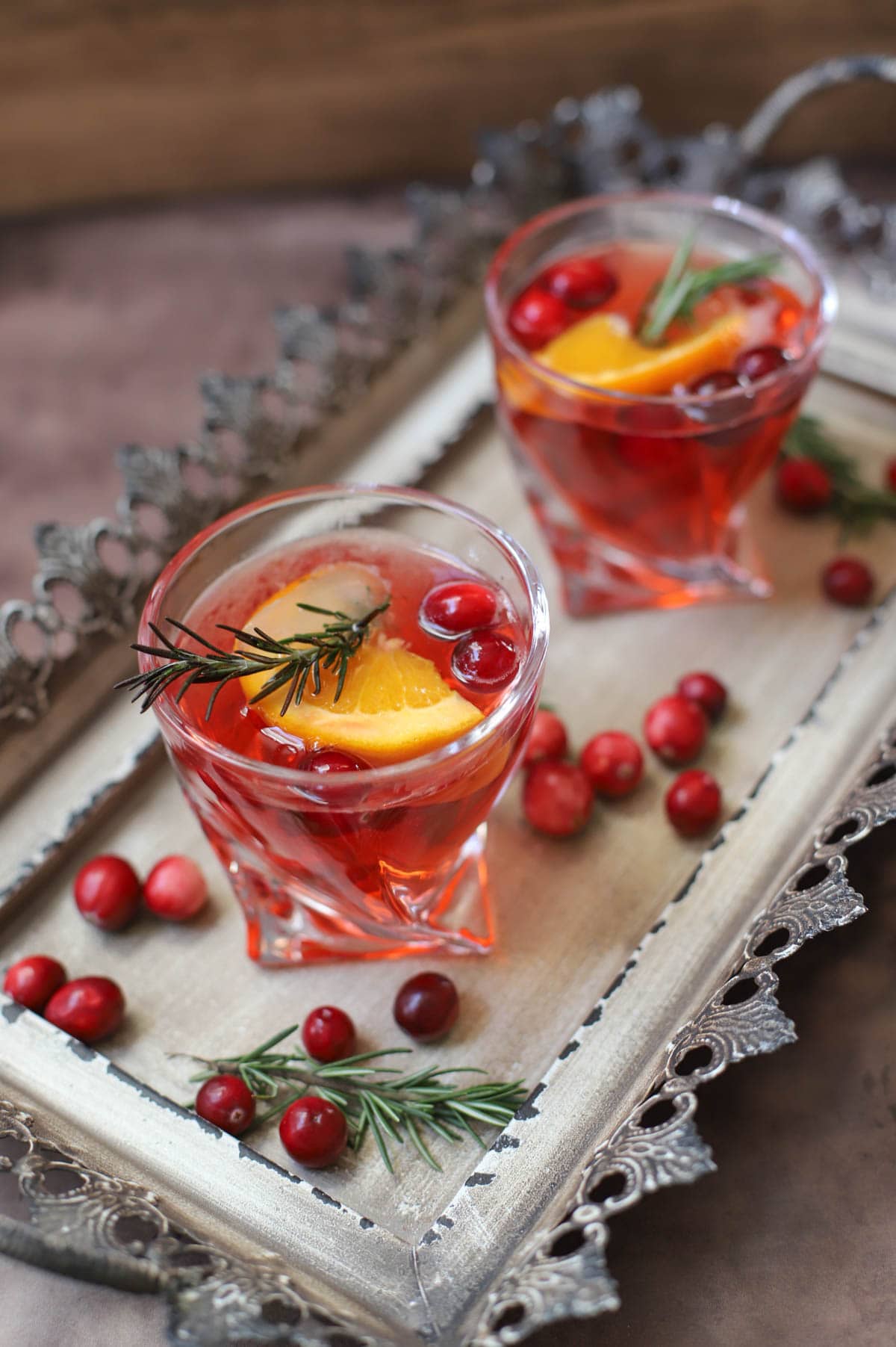 Red cocktail in a lowball glass with rosemary, cranberries, and an orange slice on a tray with cranberries and rosemary sprigs.