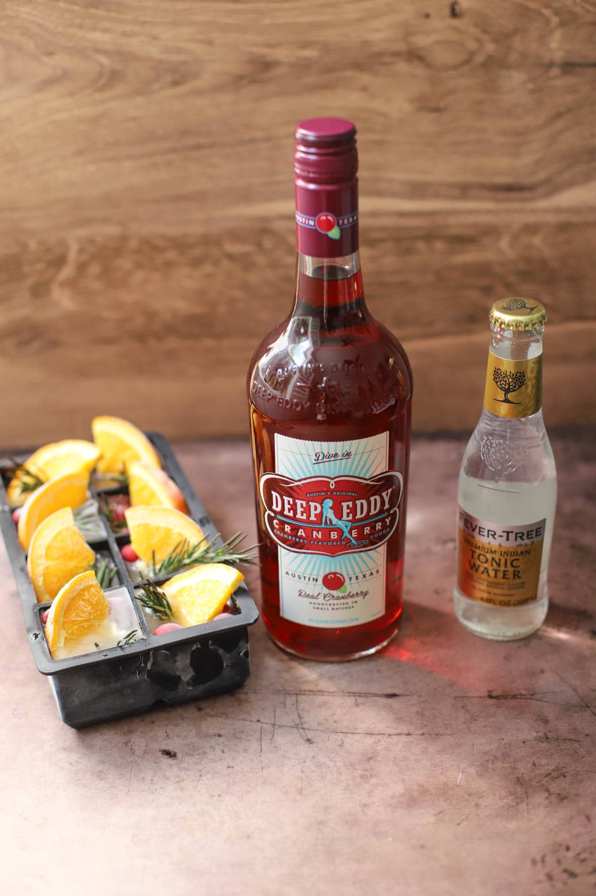 Ice cube tray filled with fruit, bottle of Deep Eddy Vodka, and bottle of tonic water on brown table.