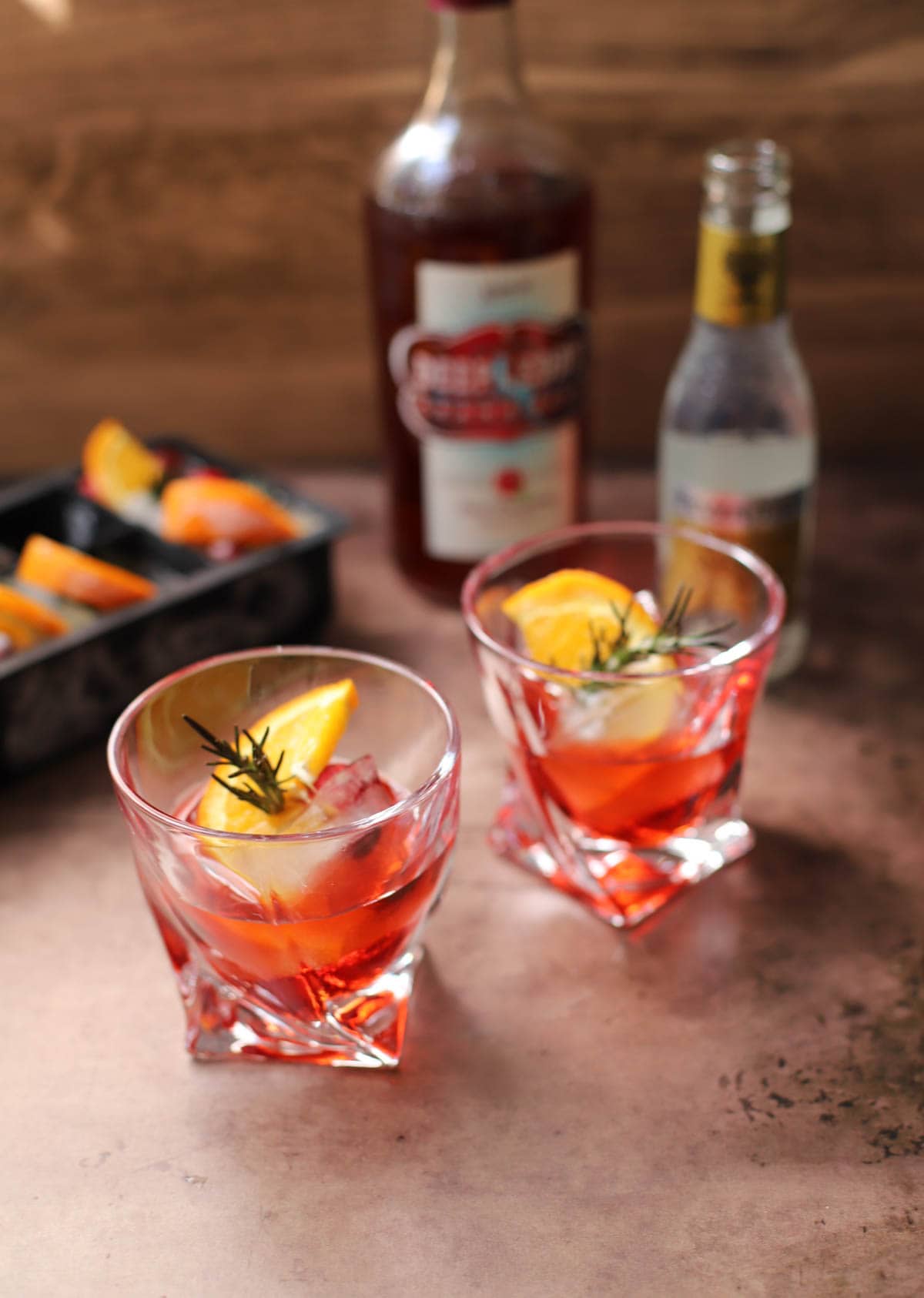 Red cocktail in a lowball glass with rosemary, cranberries, and an orange slice on brown table with bottle of red liquid, bottle of tonic, and cie cube tray in background..