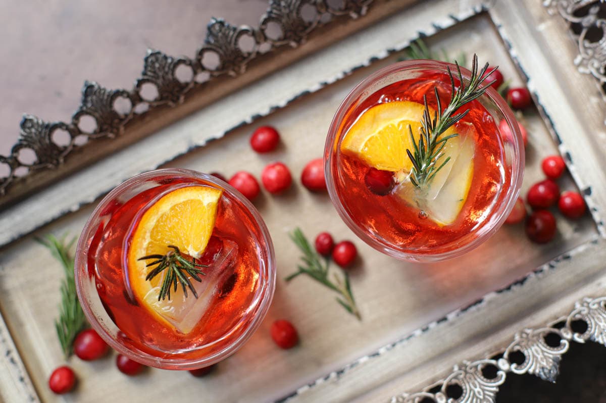 Red cocktail in a lowball glass with rosemary, cranberries, and an orange slice on a tray with cranberries and rosemary sprigs.