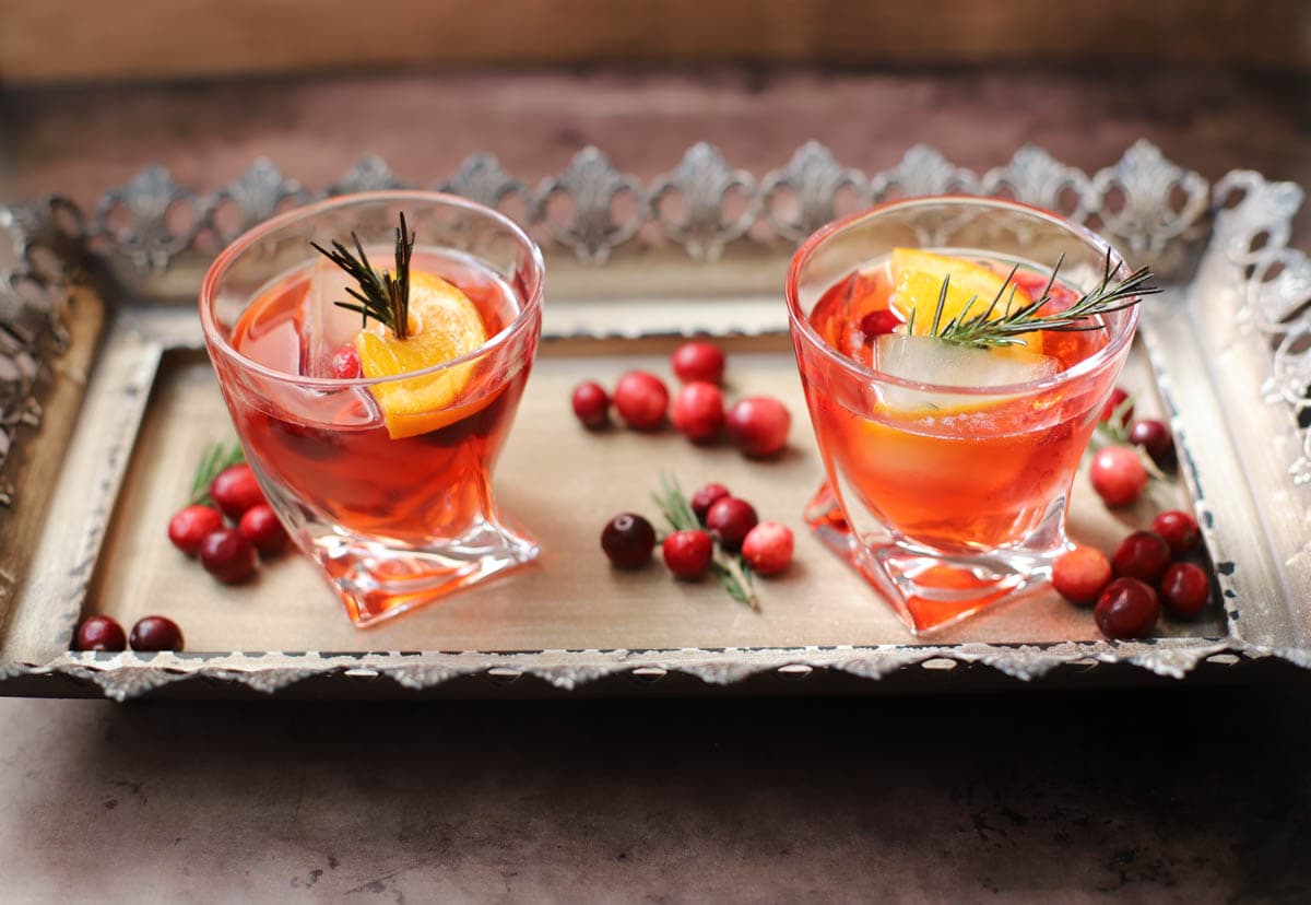 Red cocktail in a lowball glass with rosemary, cranberries, and an orange slice on a tray with cranberries and rosemary sprigs.