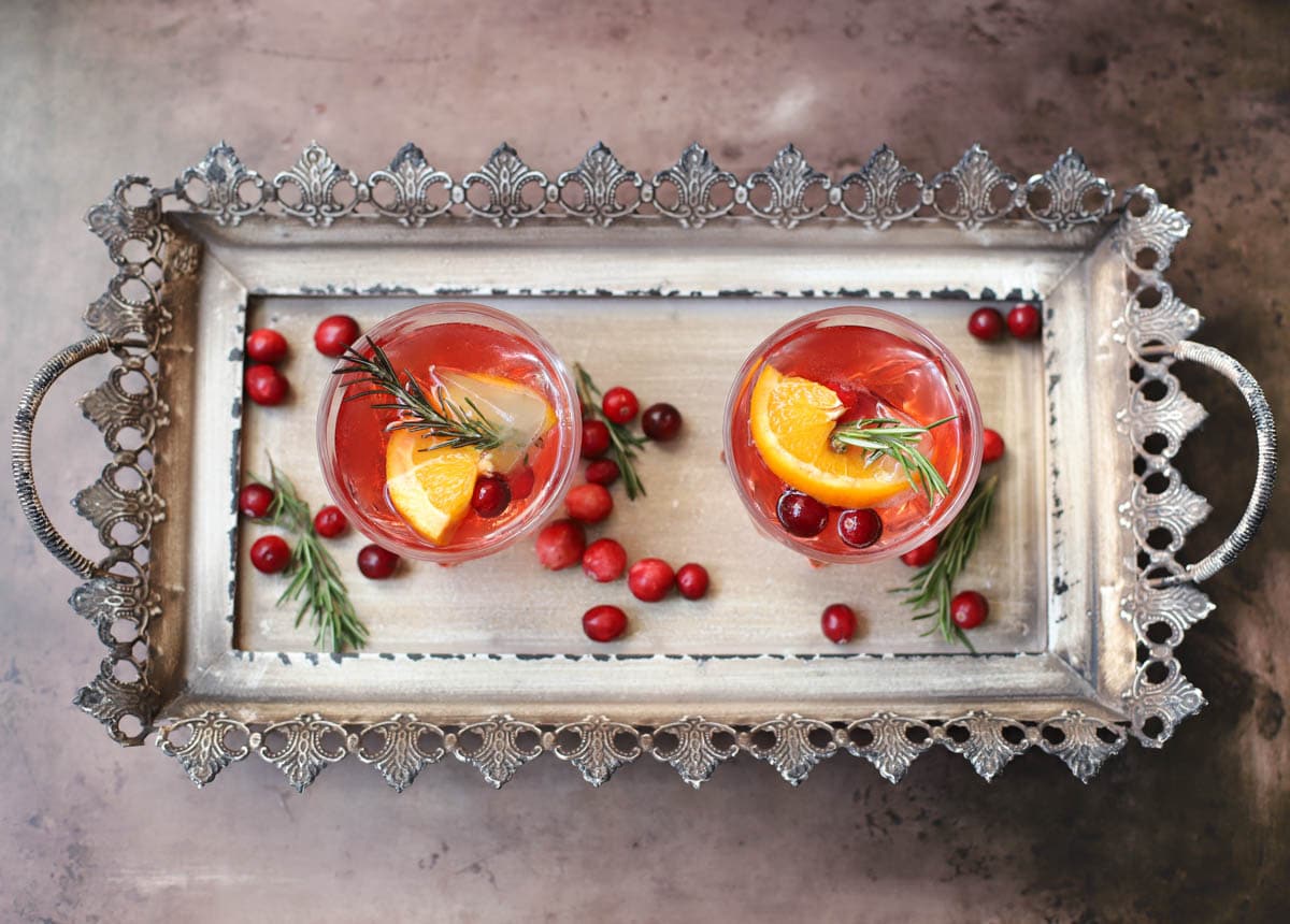 Red cocktail in a lowball glass with rosemary, cranberries, and an orange slice on a tray with cranberries and rosemary sprigs.