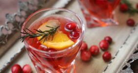 Red cocktail in a lowball glass with rosemary, cranberries, and an orange slice on a tray for Pinterest.