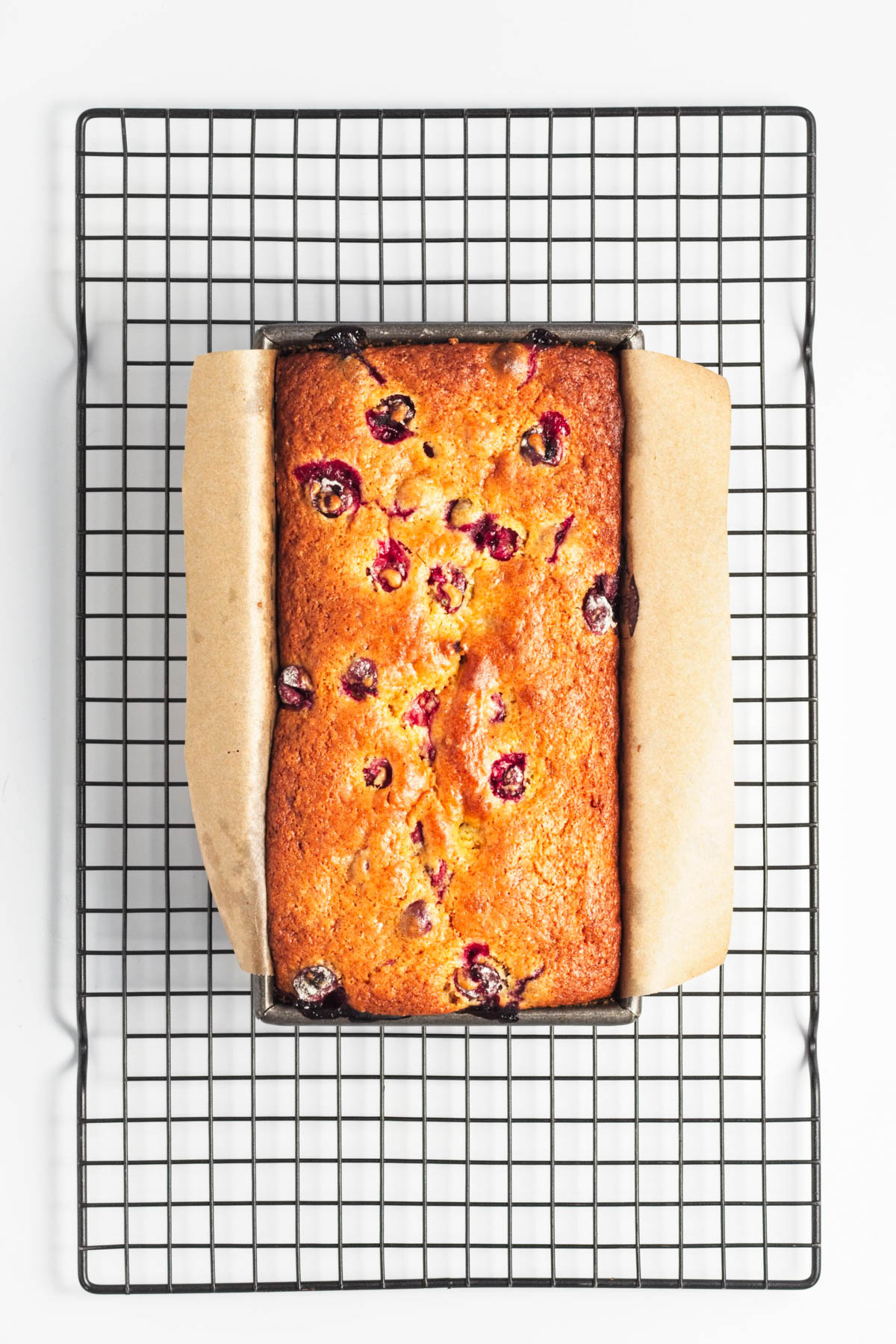 Loaf of cranberry bread in pan with parchment paper on a cooling rack.