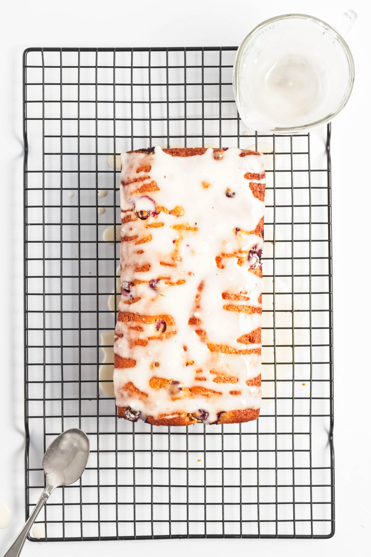 Iced loaf of cranberry bread on a cooling rack with cup of icing and a spoon.