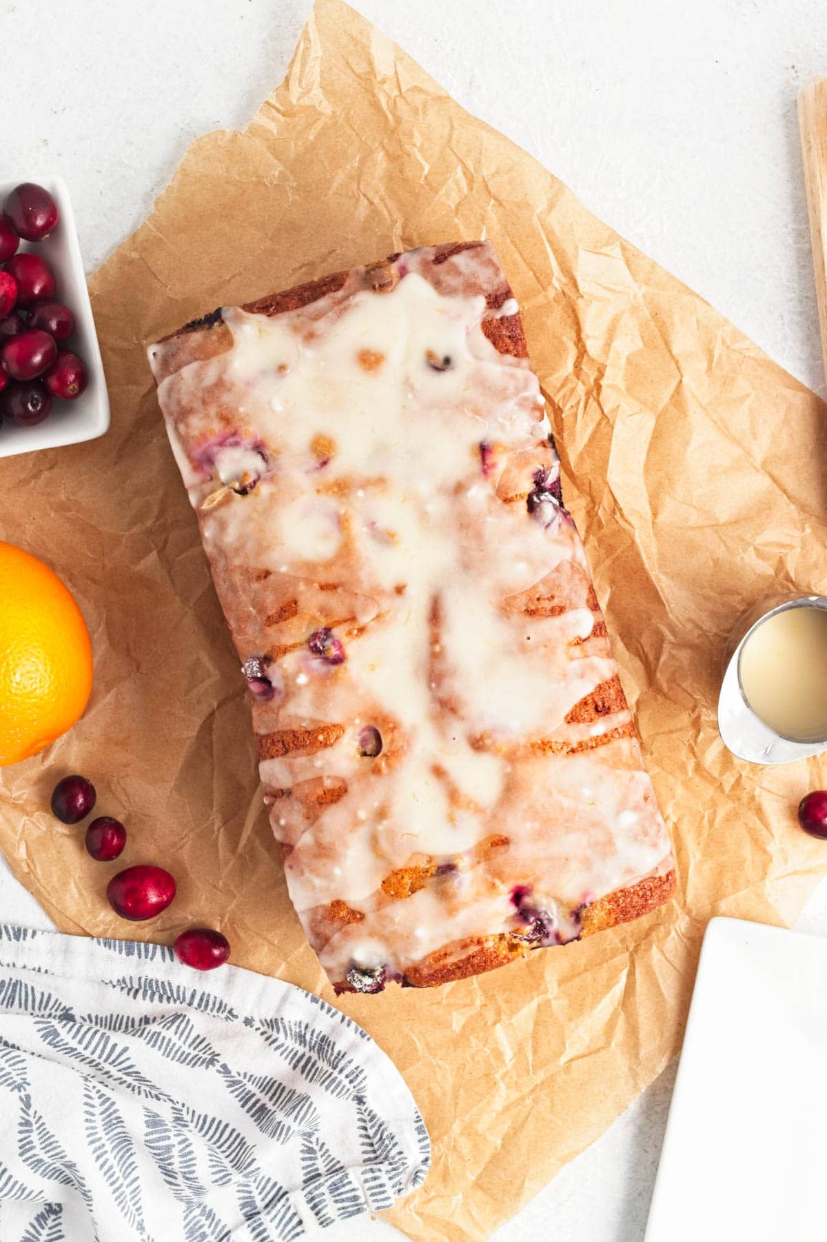 Iced cranberry bread on parchment paper with bowl of cranberries to the side along with a dish towel and small cup of icing.