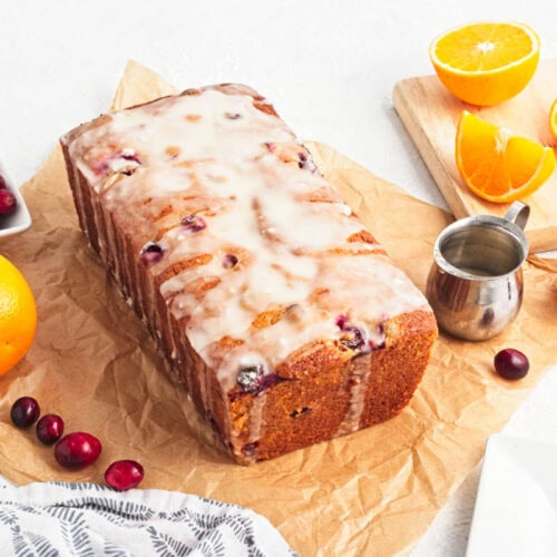 Iced cranberry bread on parchment paper with bowl of cranberries, and orange, with slices of orange on cutting board to the side along with a dish towel and small cup of icing.