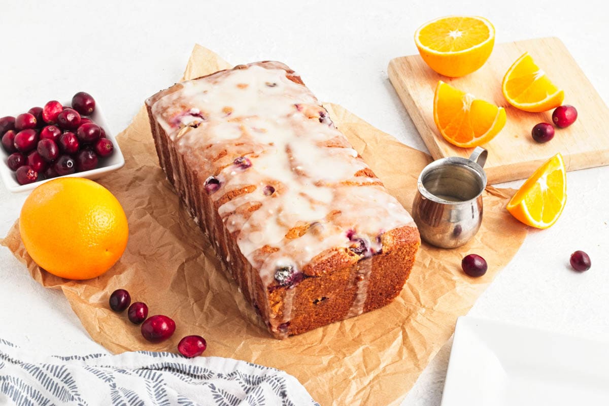 Iced cranberry bread on parchment paper with bowl of cranberries, and orange, with slices of orange on cutting board to the side along with a dish towel and small cup of icing.