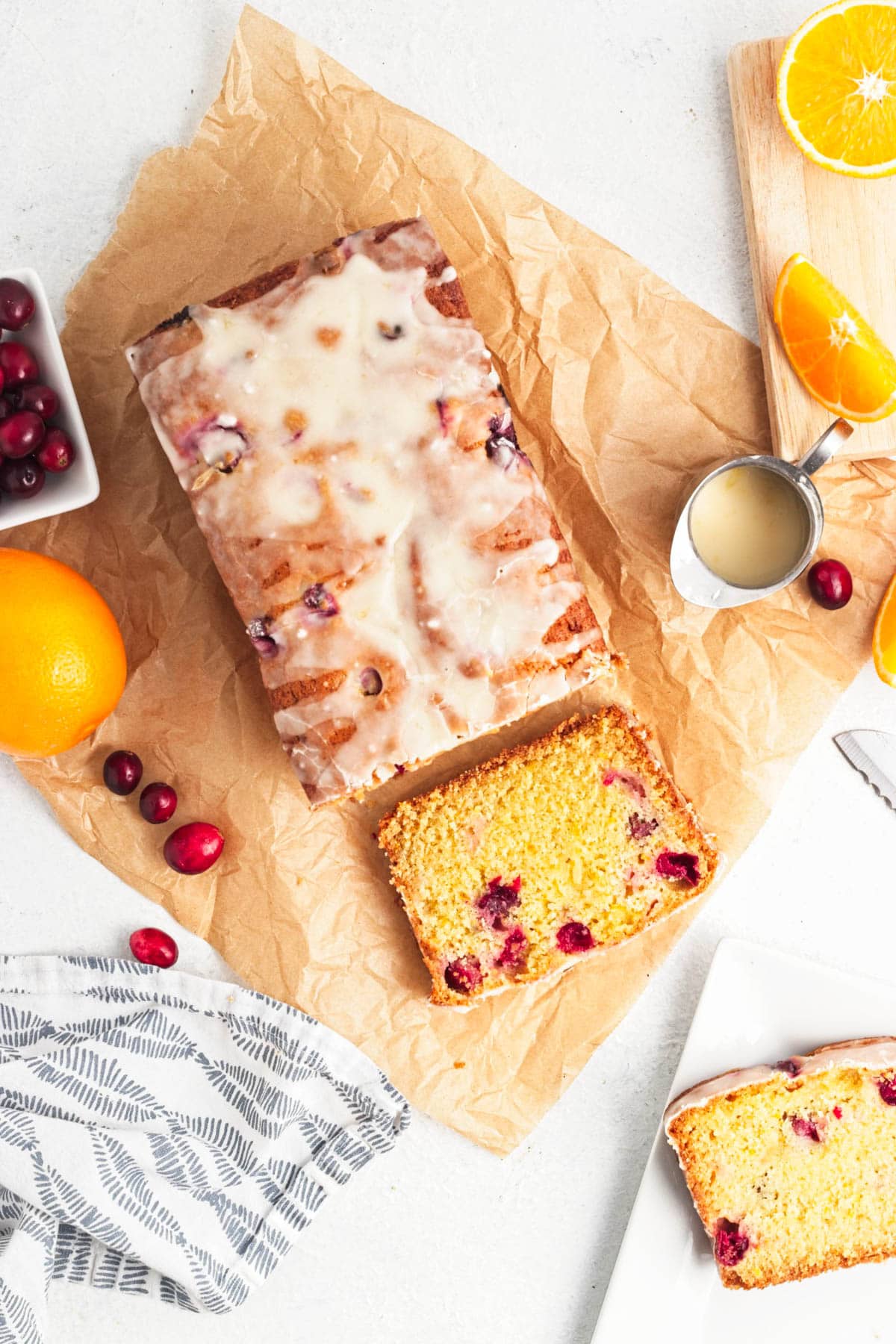 Iced cranberry bread on parchment paper with bowl of cranberries and an orange to the side along with a dish towel and small cup of icing.