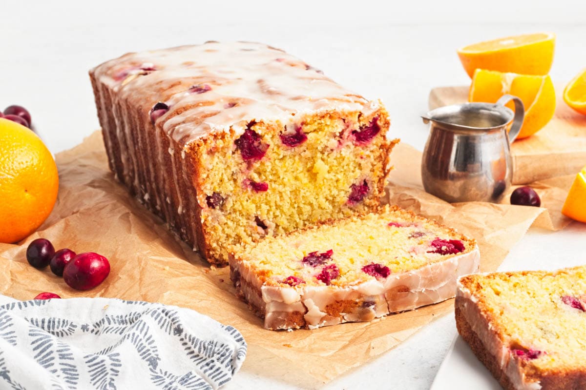 Iced cranberry bread on parchment paper with bowl of cranberries, and orange, with slices of orange on cutting board to the side along with a dish towel and small cup of icing.