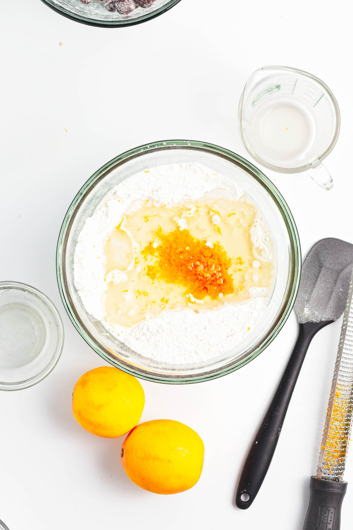 Ingredients to make cranberry orange bread in a glass bowl with a black spatula, grater, oranges, and measuring cups.