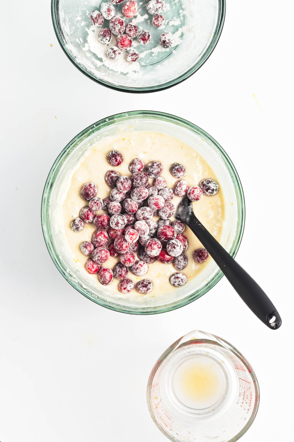 Batter with cranberries in a glass bowl with a black spatula, measuring cup, and another bowl with cranberries and flour.