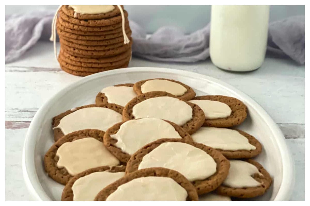 Iced dark brown cookies on a white plate with stack of cookies and jar of milk in background.