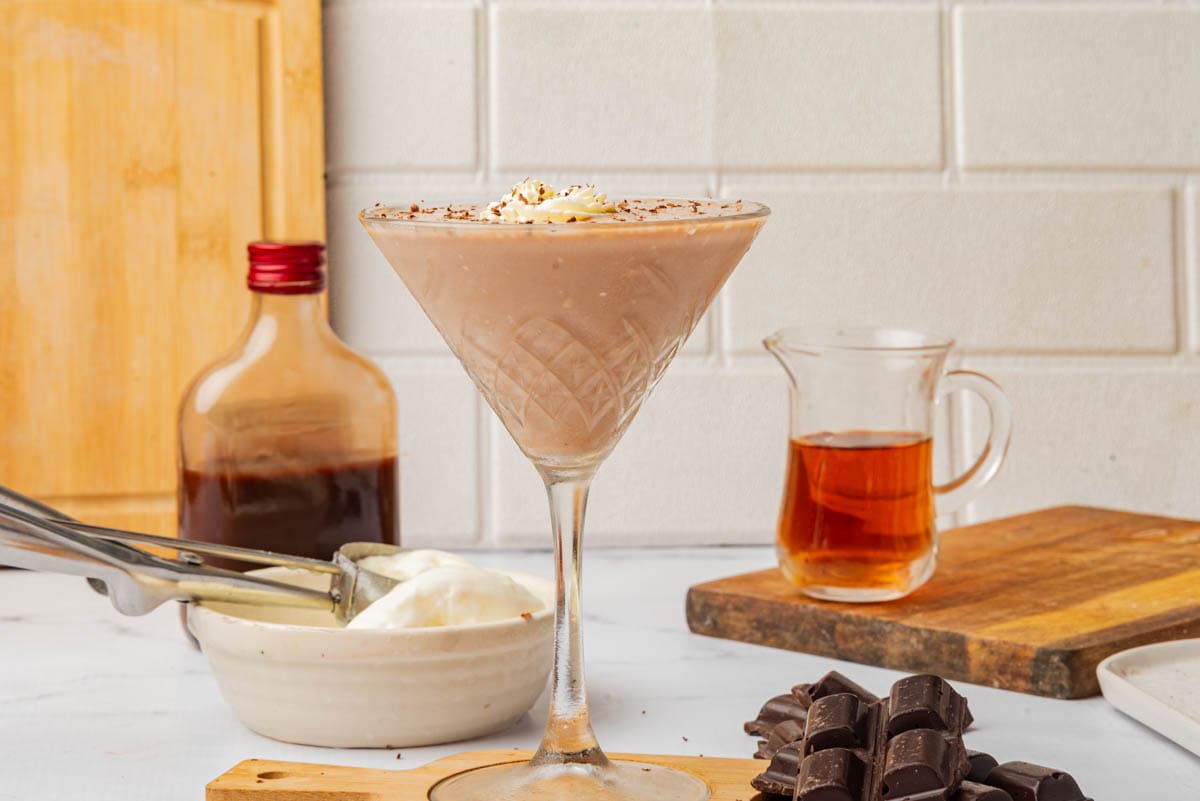 Chocolate frozen drink in a tall cocktail glass with shaved chocolate and whipped cream on a small wood cutting board with a bottle of creme de cacao, a small pitcher of brandy, a bowl of ice cream, and bars of chocolate.