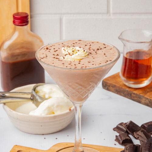 Chocolate frozen drink in a tall cocktail glass with shaved chocolate and whipped cream on a small wood cutting board with bottle of creme de cacao, a small pitcher of brandy, a bowl of ice cream, and bars of chocolate.