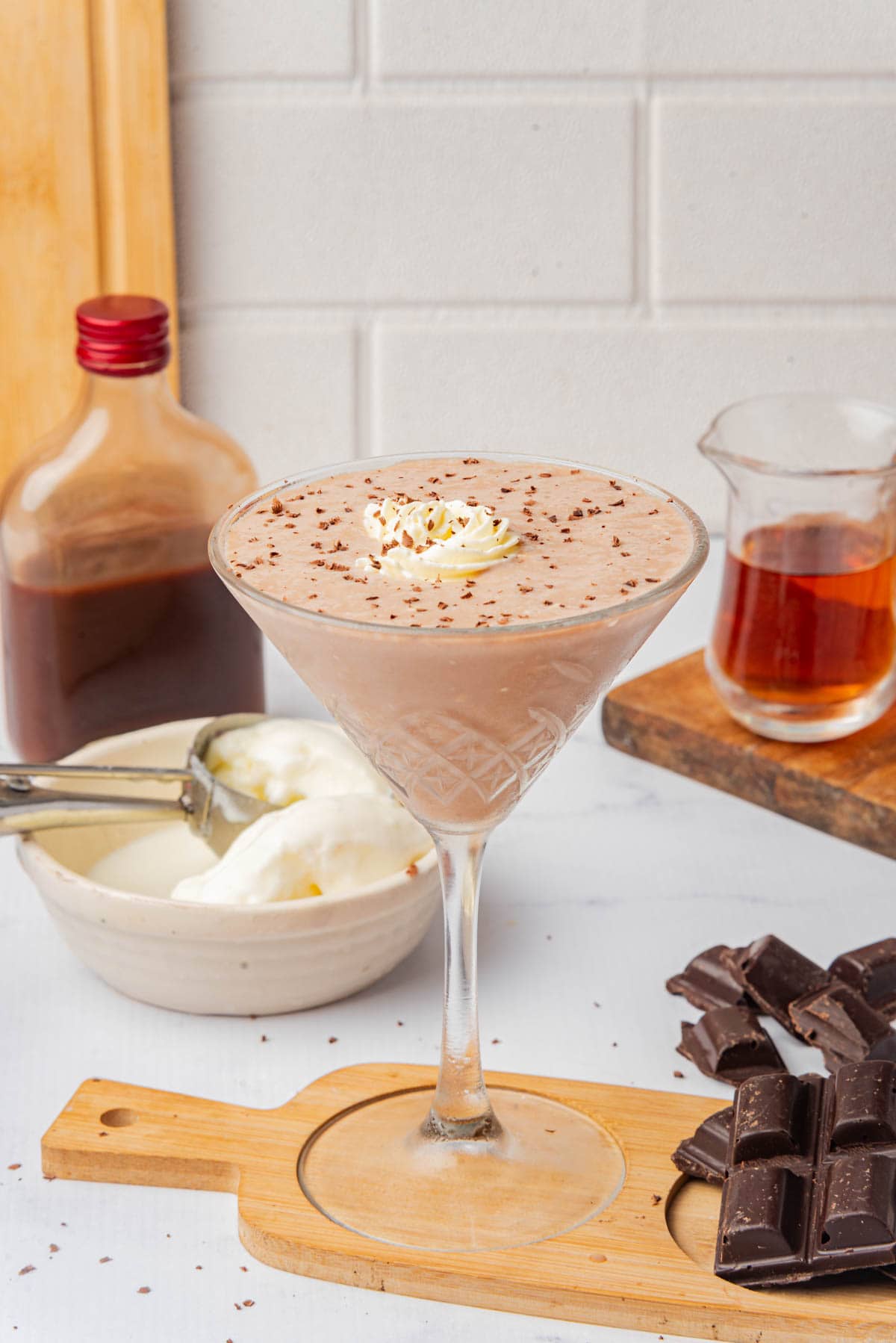 Chocolate frozen drink in a tall cocktail glass with shaved chocolate and whipped cream on a small wood cutting board with bottle of creme de cacao, a small pitcher of brandy, a bowl of ice cream, and bars of chocolate.