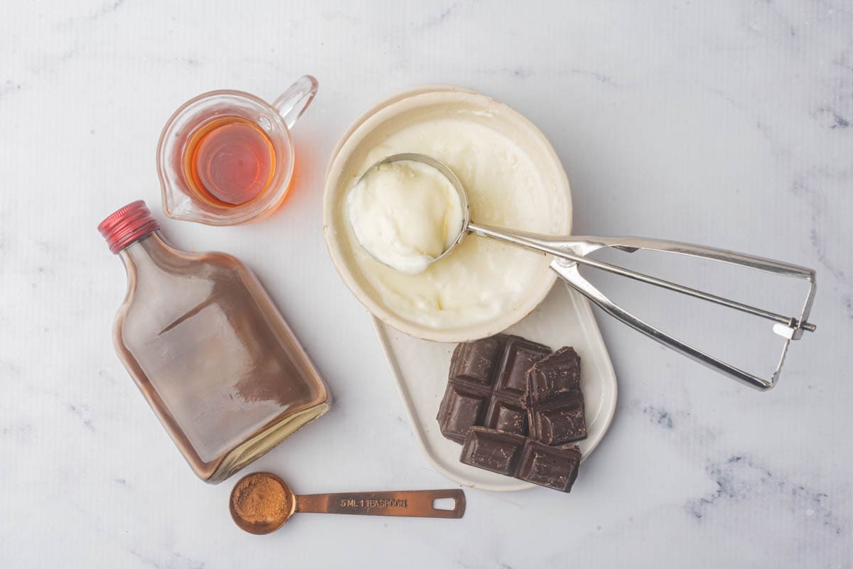 Vanilla ice cream with scoop in bowl with chocolate pieces on plate underneath with teaspoon of grated chocolate, a bottle of dark liquid, and a small pitcher of brandy.