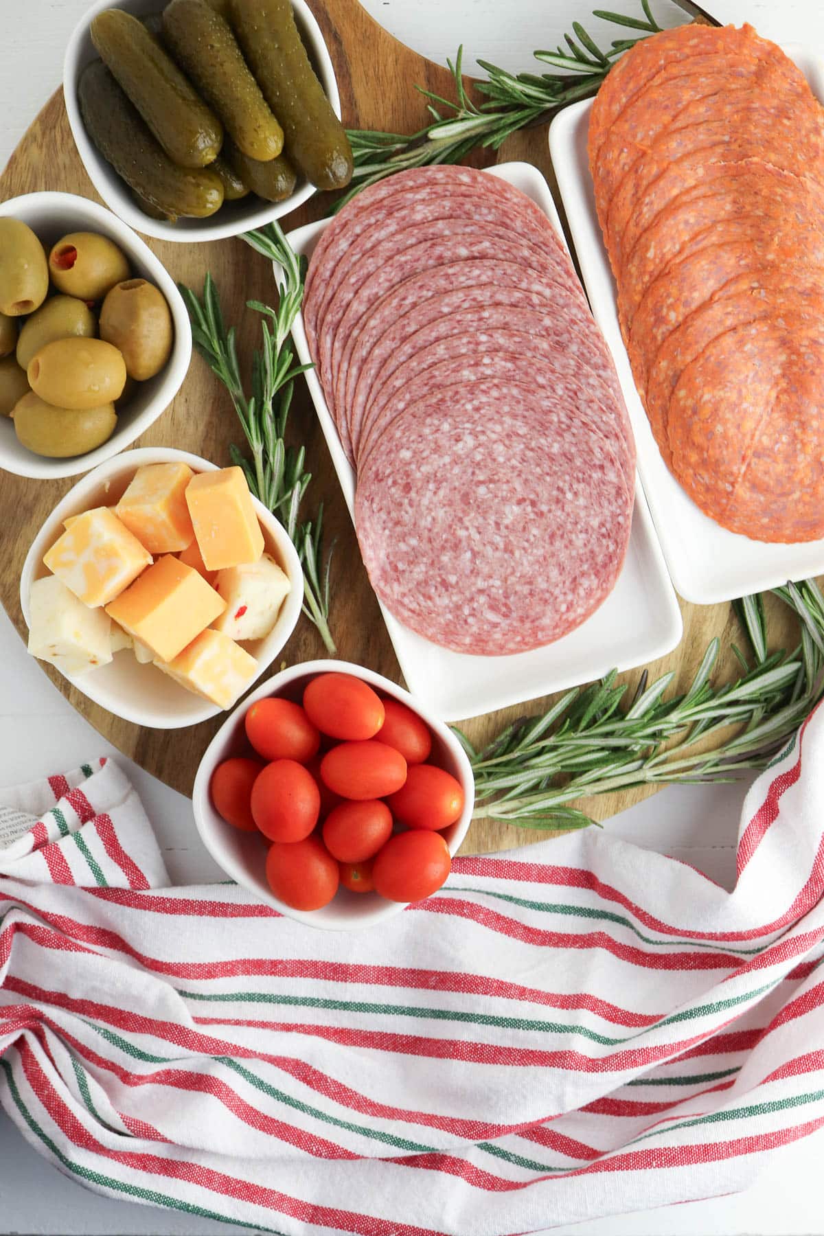 Pickles in a white bowl, olives in a white bowl, cheese in a white bowl, tomatoes in a white bowl, and salami and pepperoni on white plates with rosemary around and red, green, and white cloth.