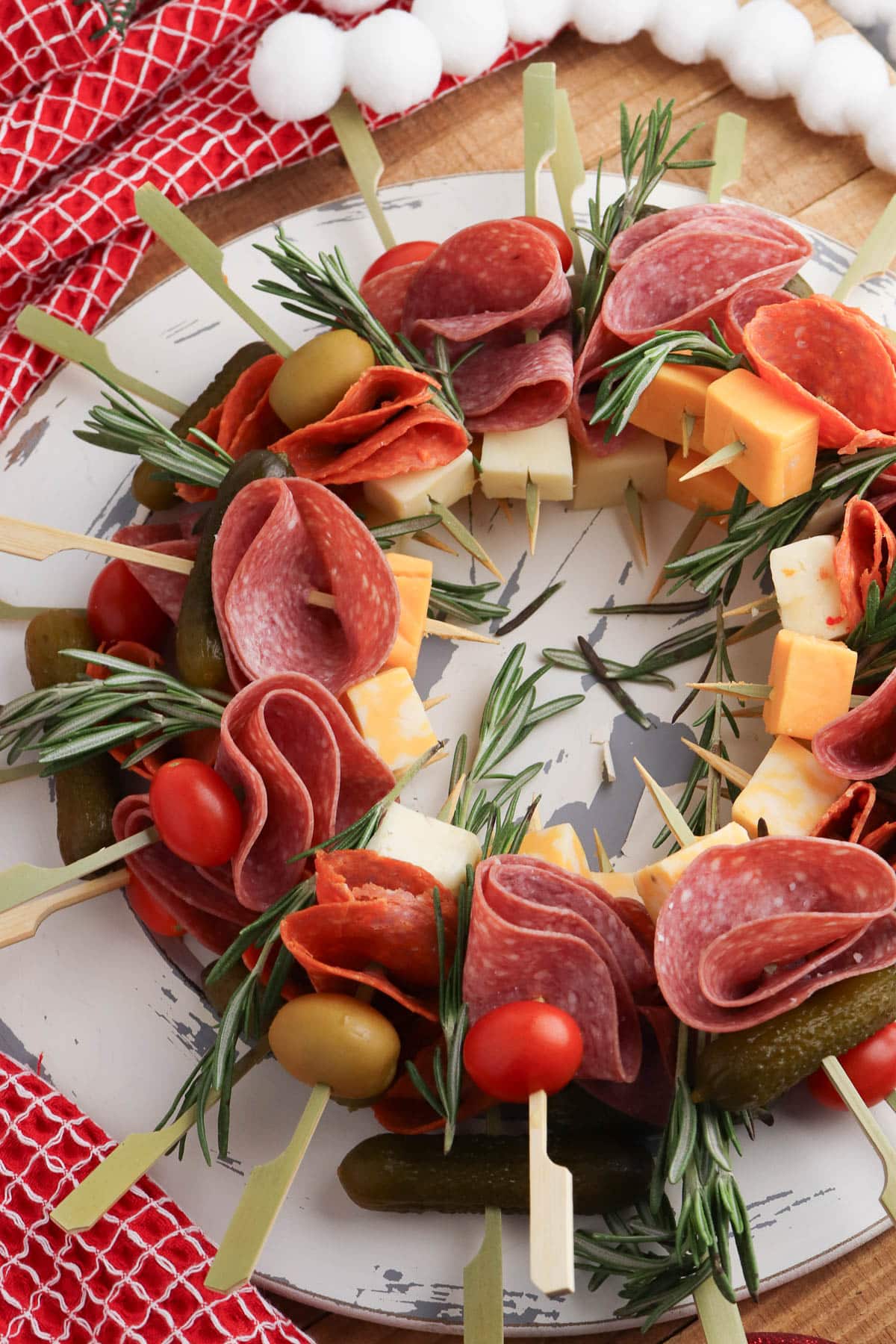 Charcuterie wreath with salami, cheese, rosemary, tomatoes, and pickles on a marble plate with red cloth and cotton balls.