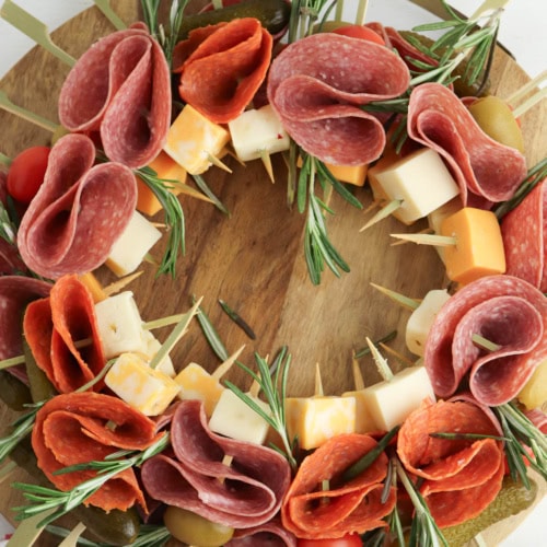 Close up of a charcuterie wreath with salami, cheese, rosemary, tomatoes, and pickles on marble plate on wood board with red ornaments.