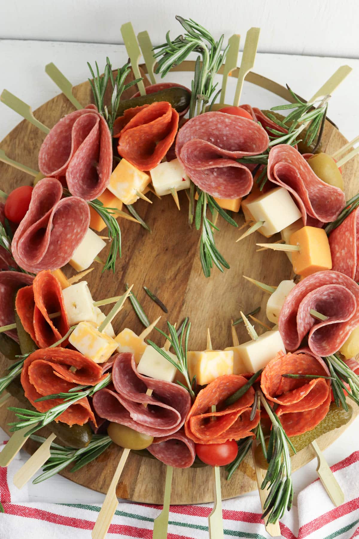 Close up of a charcuterie wreath with salami, cheese, rosemary, tomatoes, and pickles on marble plate on wood board with red ornaments.