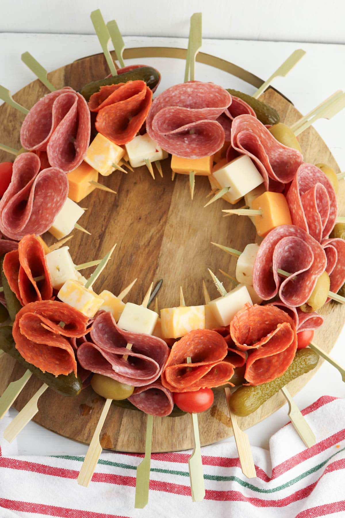 Charcuterie wreath with salami, cheese, rosemary, tomatoes, and pickles on marble plate on wood board with red ornaments.