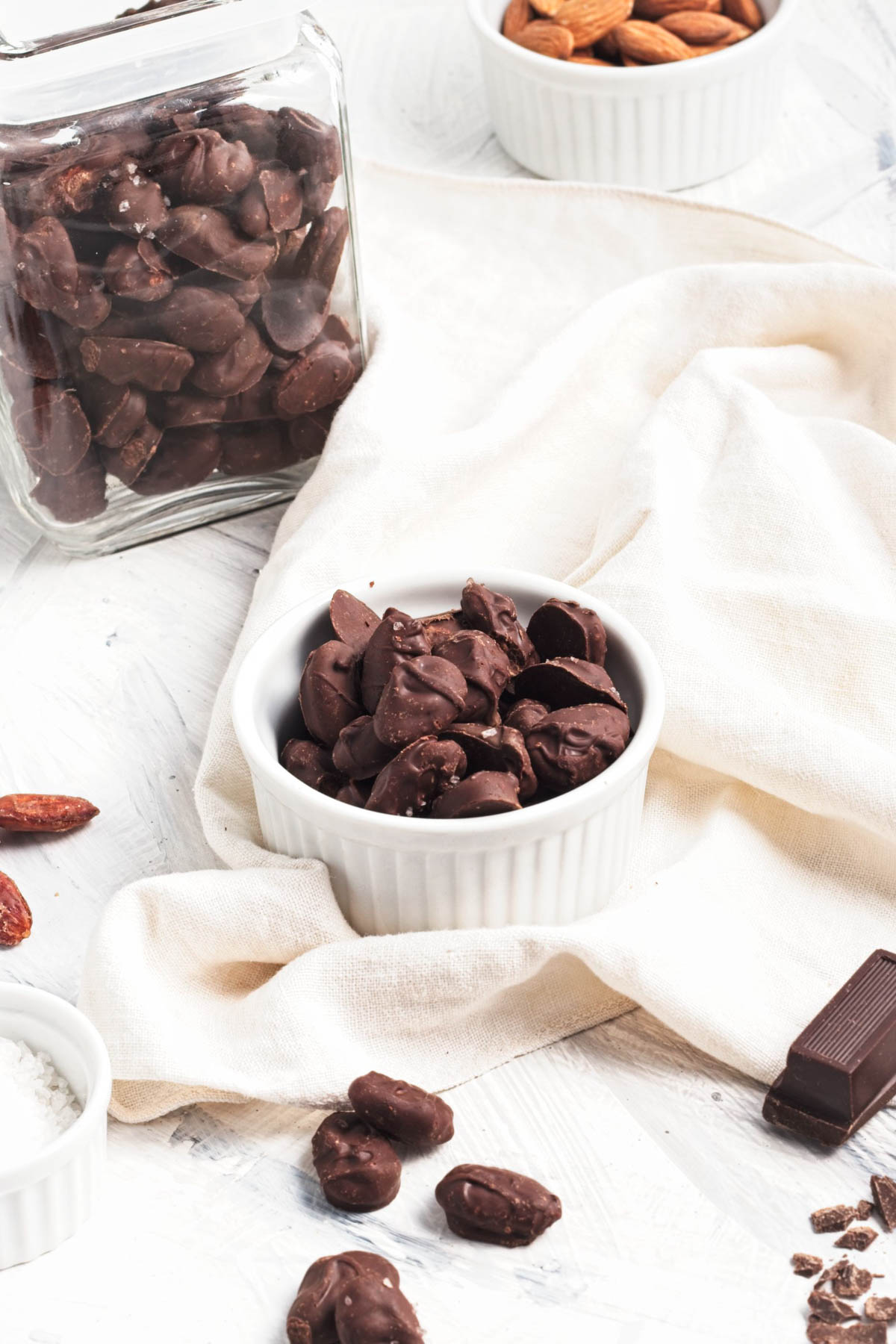 Chocolate covered almonds in a bowl on a shite napkin with a jar of more behind and a small bowl of raw almonds in background.