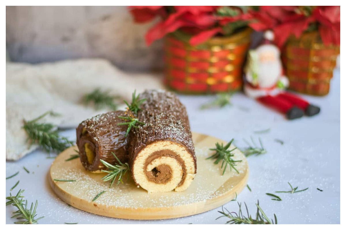 Chocolate covered white rolled cake with rosemary clippings and holiday decor in background.