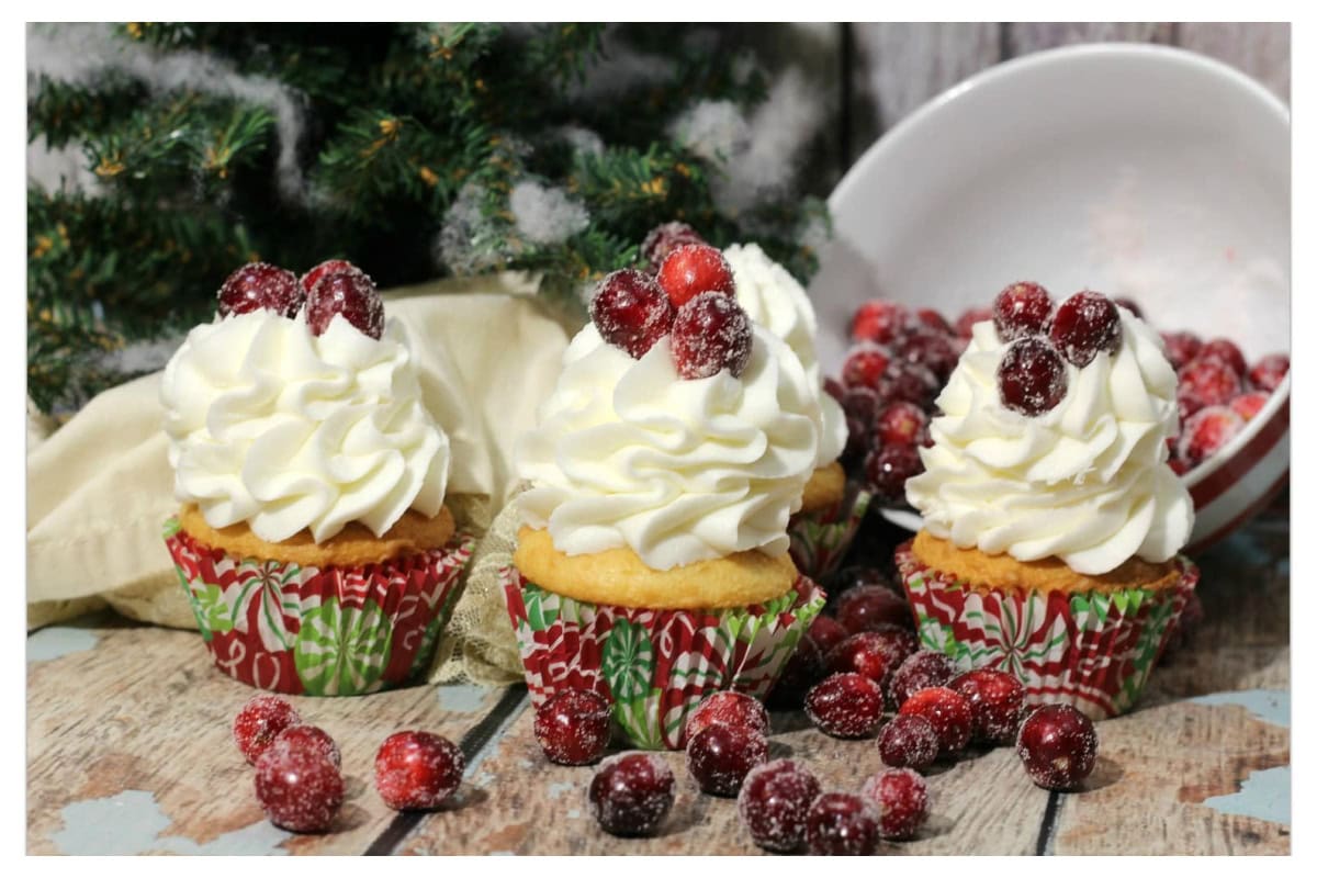 Cupcakes with cream cheese frosting and sugared cranberries on top of a wood table.