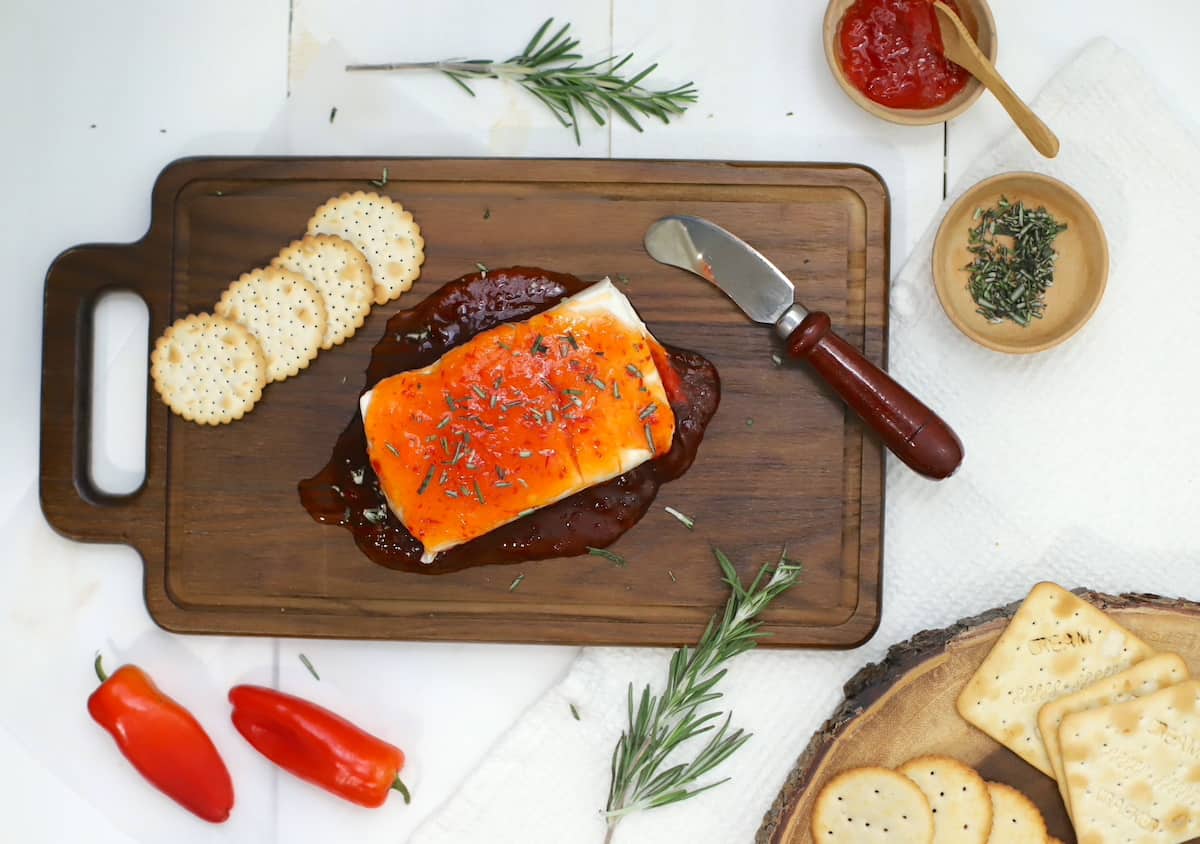 A block of cream cheese with pepper jelly poured over it with crackers and a knife on a wood board.