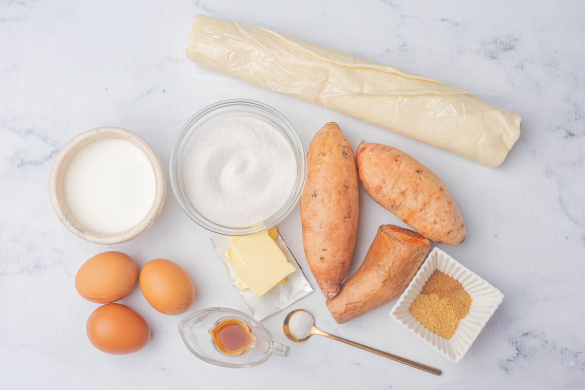 Pie crust still wrapped, sweet potatoes, spices and brown sugar in white bowl, butter, vanilla, gold spoon of sugar, eggs, sugar, milk.