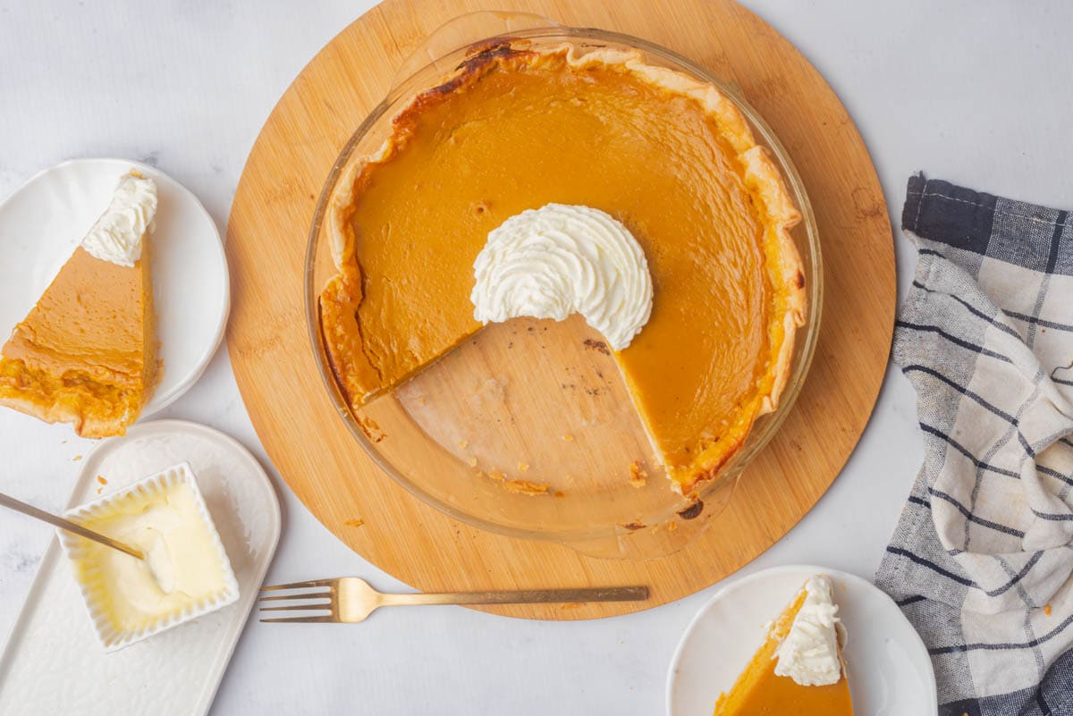 Sweet potato pie in a glass pie dish with whipped cream in center ona round wood board with fork in front, with square dish of butter to the side with dish towel and 2 pieces of pie with whipped cream.