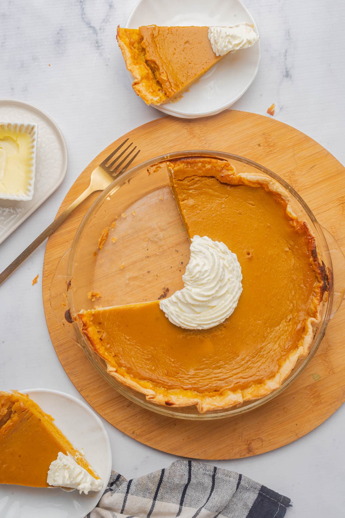 Sweet potato pie in a glass pie dish with whipped cream in center, square dish of butter to the side with dish towel and 2 pieces of pie with fork on side of wood board.