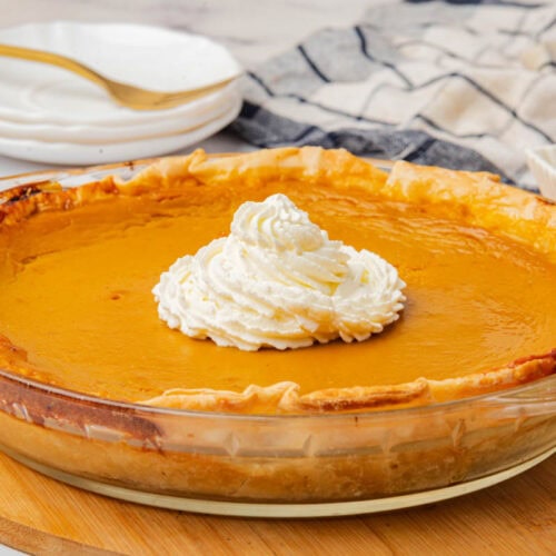 Sweet potato pie in a glass pie dish with whipped cream in center, square dish of butter in background with dish towel and 3 saucers with fork.