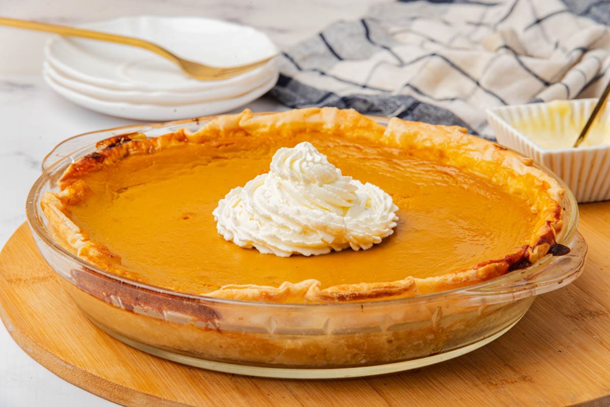 Sweet potato pie in a glass pie dish with whipped cream in center, square dish of butter in background with dish towel and 3 saucers with fork.