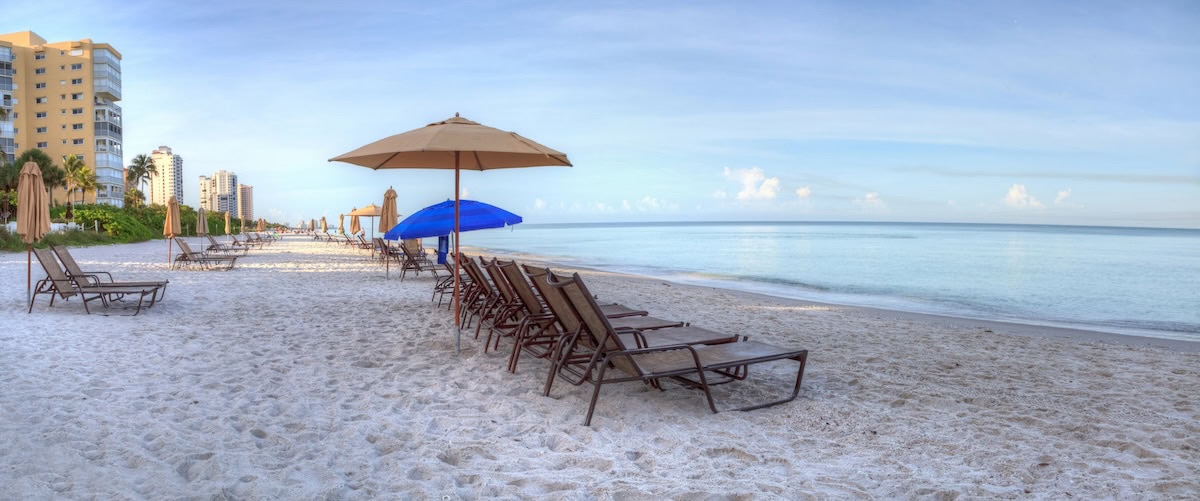 Umbrellas and chairs on beach at sunrise with a calm ocean and buildings along the white sand.