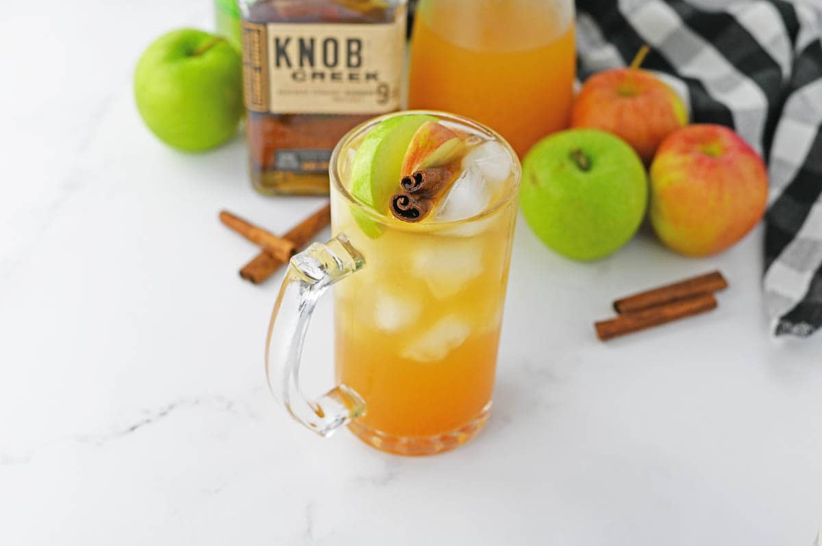 Glass of apple cider with cinnamon stick and apple slices with bourbon bottle and apples and ginger ale in background.