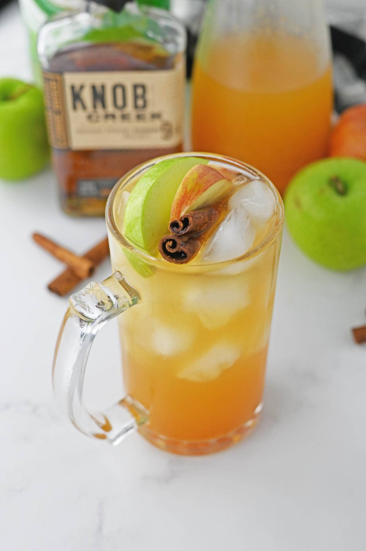 Glass of apple cider with cinnamon stick and apple slices with bourbon bottle and apples and ginger ale in background.