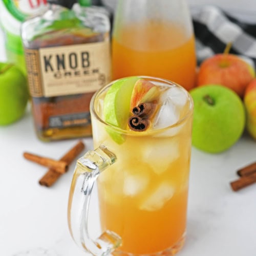 Glass of apple cider with cinnamon stick and apple slices with bourbon bottle and apples and ginger ale in background.