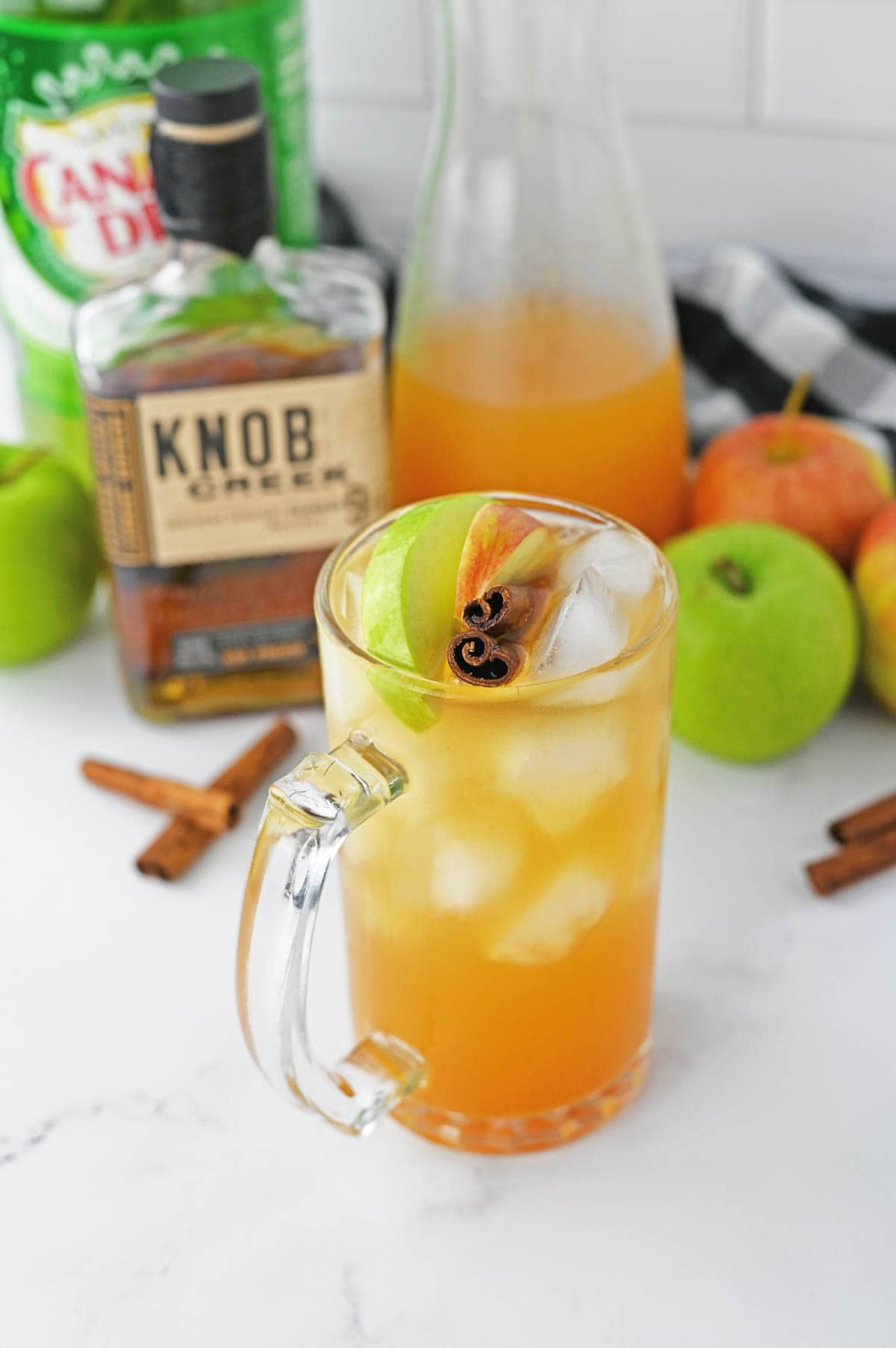 Glass of apple cider with cinnamon stick and apple slices with bourbon bottle and apples and ginger ale in background.